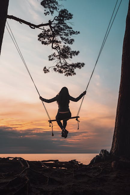 pexels-photo-4300115-4300115 A woman swings on a tree above the sea at sunset, creating a serene silhouette.